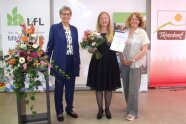 2 Frauen stehen nebeneinander, eine hält Blumenstrauß und Urkunde